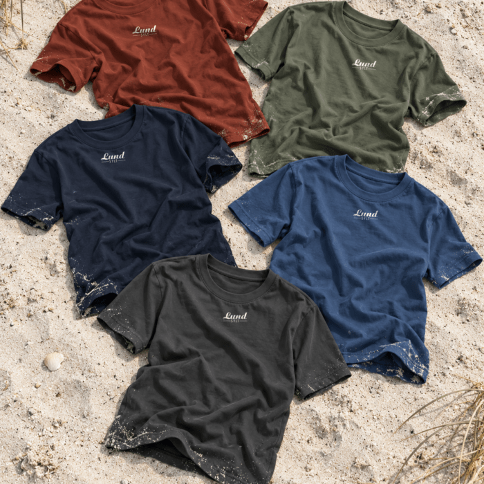 Fünf Sylt T-Shirts von Lund Sylt mit weißem „Lund Sylt“-Logo in Erdrot, Aloe, Navy, Arbeiterblau und Anthrazit liegen locker verteilt auf hellem Sylter Strandsand (Bild mit KI erstellt)