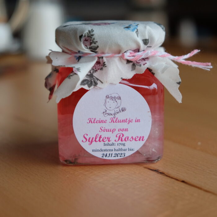 DSCF0064 Ein Glas Rosensirup mit geblümtem Stoffdeckel steht auf einem Holztisch neben einer Schale mit SYLTER ROSEN-KANDIS „KLUNTJES“.