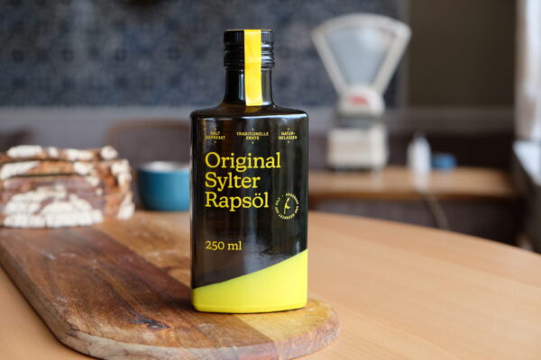 DSCF0093 Eine Flasche SYLTER RAPSÖL Kaltgepresst, nativ, 250 ml, wird elegant auf einem Holzbrett präsentiert, im Hintergrund Brot und eine Waage. Dieses kaltgepresste Öl verleiht Ihrem kulinarischen Erlebnis einen Hauch von Reinheit und Tradition.