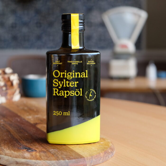 DSCF0093 Eine Flasche SYLTER RAPSÖL Kaltgepresst, nativ, 250 ml, wird elegant auf einem Holzbrett präsentiert, im Hintergrund Brot und eine Waage. Dieses kaltgepresste Öl verleiht Ihrem kulinarischen Erlebnis einen Hauch von Reinheit und Tradition.