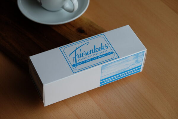 Friesenkekse von Lund Sylt Eine Schachtel FRIESENKEKSE 240 g steht neben einer weißen Teetasse und Untertasse auf einem Holztisch.