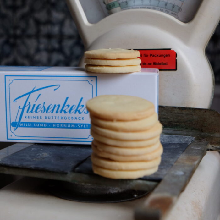 Ein Stapel FriesenKekse Ein Stapel FriesenKekse balanciert auf einer Vintage-Waage, im Hintergrund steht eine Schachtel mit der Aufschrift „FRIESENKEKSE 240 g“.