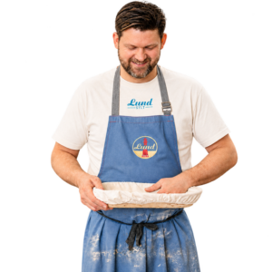 Ein Mann in einer blauen Schürze und dem Lund-Shirt – mit blauem Logo – hält eine abgedeckte runde Auflaufform in der Hand, lächelt und blickt darauf hinunter.