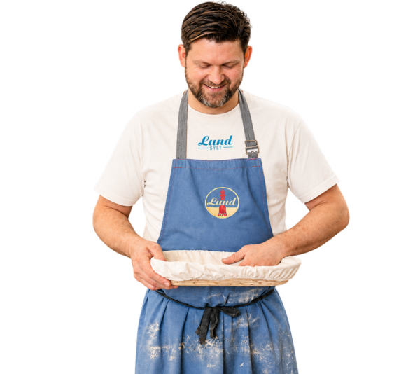 Ein Mann in einer blauen Schürze und dem Lund-Shirt – mit blauem Logo – hält eine abgedeckte runde Auflaufform in der Hand, lächelt und blickt darauf hinunter.
