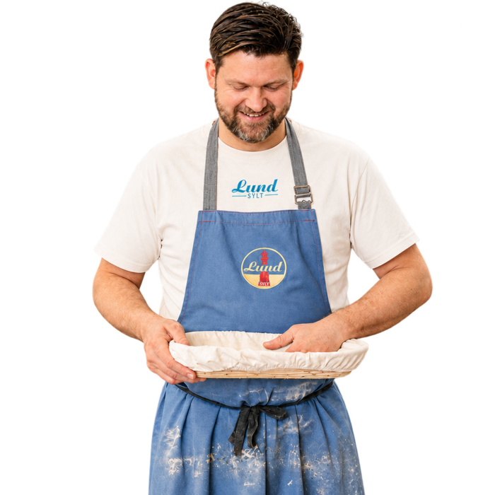 Ein Mann in einer blauen Schürze und dem Lund-Shirt – mit blauem Logo – hält eine abgedeckte runde Auflaufform in der Hand, lächelt und blickt darauf hinunter.