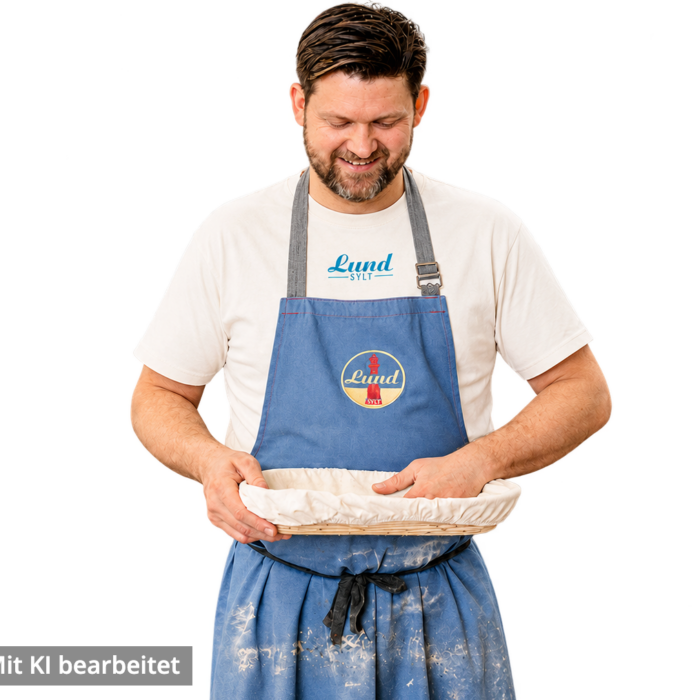 Ein Mann in einer blauen Schürze und dem Lund-Shirt – mit blauem Logo – hält eine abgedeckte runde Auflaufform in der Hand, lächelt und blickt darauf hinunter (Bild mit KI bearbeitet)