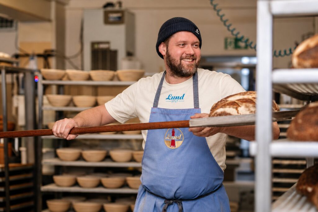Jens Lund in Lund-Shirt und Lund-Schürze holt Brot aus dem Ofen der Backstube in Hörnum