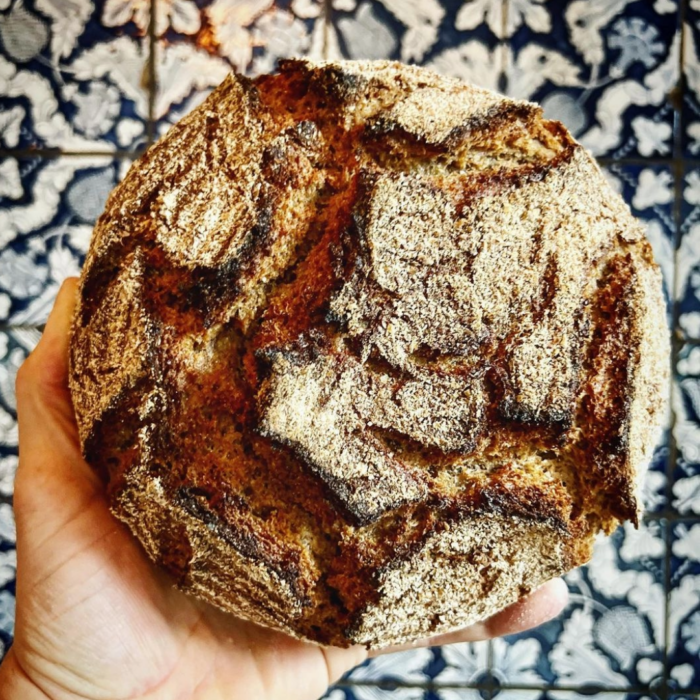 Lund Sylt Bio SYLTER Roggen und Dinkel back Hand hält rundes handwerklich hergestelltes Brot mit rustikaler Kruste vor einem gemusterten blau-weißen Fliesenhintergrund.