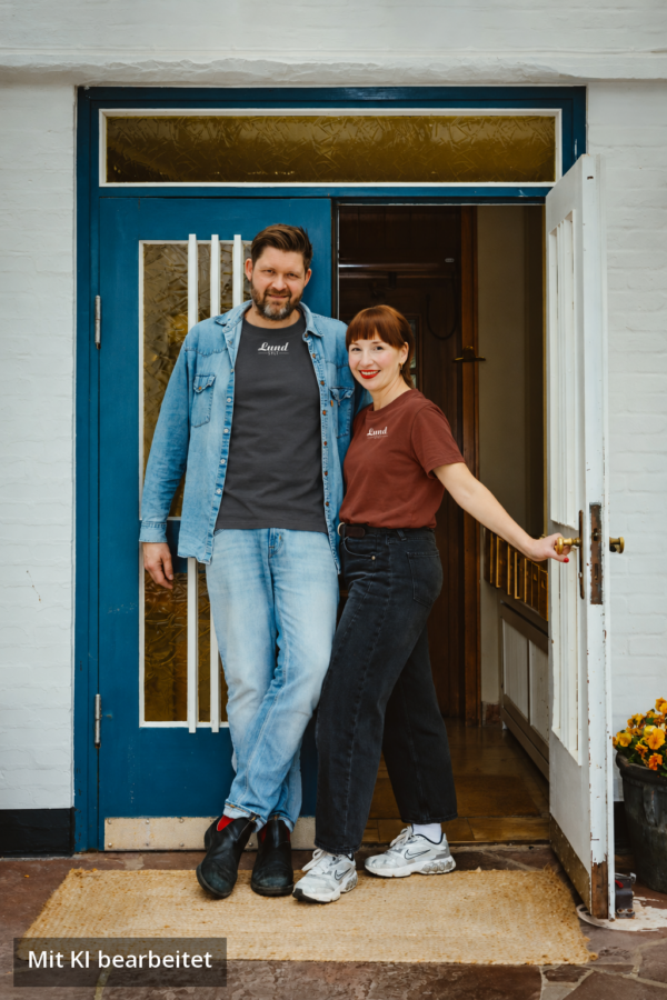 Lund Workwear: Jens Lund und Julia im Sylt T-Shirt mit weißem Logo (Anthrazit und Erdrot) Jens Lund und Julia von Lund Sylt stehen vor einer blauen Holztür und tragen das LUND SHIRT mit weißem „Lund Sylt“-Logo, er in Anthrazit unter einer offenen Jeansjacke, sie in Erdrot (Bild mit KI bearbeitet)