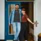 Jens Lund und Julia von Lund Sylt stehen vor einer blauen Holztür und tragen das LUND SHIRT mit weißem „Lund Sylt“-Logo, er in Anthrazit unter einer offenen Jeansjacke, sie in Erdrot (Bild mit KI bearbeitet)