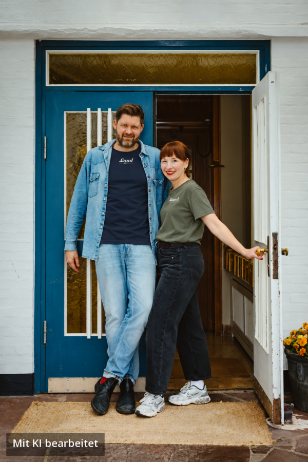 Lund Workwear: Jens Lund und Julia im Sylt T-Shirt mit weißem Logo (Navy und Aloe) Jens Lund und Julia von Lund Sylt stehen vor einer blauen Holztür und tragen das LUND SHIRT mit weißem „Lund Sylt“-Logo, er in Navy unter einer offenen Jeansjacke, sie in Aloe (Bild mit KI bearbeitet)