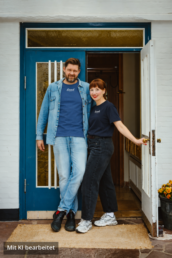 Lund Workwear: Jens Lund und Julia im Sylt T-Shirt mit weißem Logo (Arbeiterblau und Navy) Jens Lund und Julia von Lund Sylt stehen vor einer blauen Holztür und tragen das LUND SHIRT mit weißem „Lund Sylt“-Logo, er in Arbeiterblau unter einer offenen Jeansjacke, sie in Navy (Bild mit KI bearbeitet)