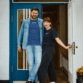 Jens Lund und Julia von Lund Sylt stehen vor einer blauen Holztür und tragen das LUND SHIRT mit weißem „Lund Sylt“-Logo, er in Arbeiterblau unter einer offenen Jeansjacke, sie in Navy (Bild mit KI bearbeitet)