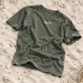 Sylt T-Shirt von Lund Sylt in Aloe (Aloe) mit weißem „Lund Sylt“-Logo auf der Brust, locker auf hellem Sylter Strandsand mit Strandhafer liegend (Bild mit KI erstellt)