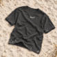 Sylt T-Shirt von Lund Sylt in Anthrazit (Anthracite) mit weißem „Lund Sylt“-Logo auf der Brust, locker auf hellem Sylter Strandsand mit Strandhafer liegend (Bild mit KI erstellt)