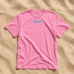 Pinkfarbenes LundSylt T-Shirt in XXL mit "Lund SYLT" in blauer Schrift auf sandfarbenem Hintergrund (Bild mit KI erstellt)
