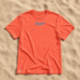 Rotoranges Lund-Shirt mit blauem Logo auf Sand (Bild mit KI erstellt)