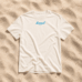 Ein weißes „LundSylt“-T-Shirt in Größe XXL mit auffälliger blauer Aufschrift liegt im strahlenden Sonnenlicht auf strukturiertem Sand.