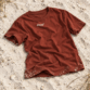 Sylt T-Shirt von Lund Sylt in Erdrot (Red Earth) mit weißem „Lund Sylt“-Logo auf der Brust, locker auf hellem Sylter Strandsand mit Strandhafer liegend (Bild mit KI erstellt)
