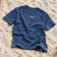 Sylt T-Shirt von Lund Sylt in Arbeiterblau (Worker Blue) mit weißem „Lund Sylt“-Logo auf der Brust, locker auf hellem Sylter Strandsand mit Strandhafer liegend (Bild mit KI erstellt)
