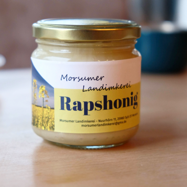 SylterRapshonig Ein Glas „Sylter Rapshonig“ mit sonnig gelbem Etikett steht einladend auf einem Holztisch und erinnert an die lebendige Sylter Landschaft.