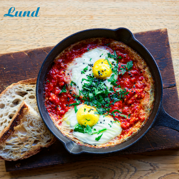 Tofreeshakshukalund Eine Pfanne TOFREE NORTH SHAKSHUKA, garniert mit Eiern und Kräutern, wird elegant mit knusprigen Brotscheiben auf einer rustikalen Holzoberfläche präsentiert.