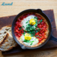 Eine Pfanne TOFREE NORTH SHAKSHUKA, garniert mit Eiern und Kräutern, wird elegant mit knusprigen Brotscheiben auf einer rustikalen Holzoberfläche präsentiert.