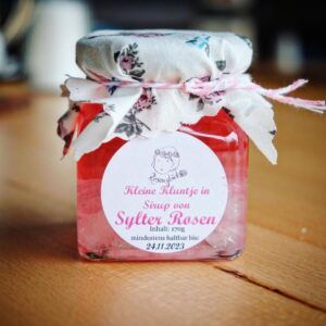 Ein Glas rosa SYLTER ROSEN-KANDIS „KLUNTJES“ Marmelade auf einem Holztisch.